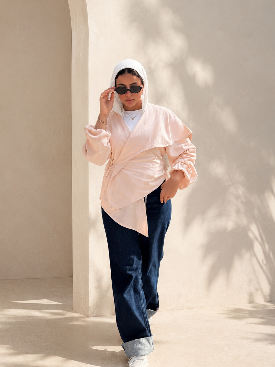 A woman wearing a light peach asymmetrical wrap blouse, dark navy wide-leg pants, a white headscarf, and white sneakers walks in a sunlit architectural setting with beige walls and leaf shadows.